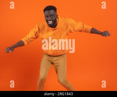 Jeune homme afro-américain excité avec de courts cheveux bouclés regardant la caméra tout en se tenant debout avec les jambes pliées bras tendus avec des poings fermés et dansant Banque D'Images