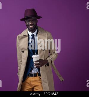 Jeune homme afro-américain souriant dans une cravate élégante et un chapeau regardant la caméra tout en se tenant debout avec une tasse à emporter près de la toile de fond violette Banque D'Images