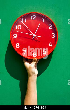 Recadrez la main d'une personne méconnaissable tenant une grande horloge rouge ronde contre le mur vert Banque D'Images