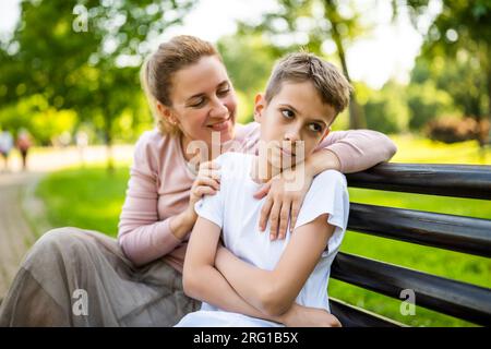 Mère heureuse est assise avec son fils sur le banc dans le parc. Garçon est offensé et boude. Banque D'Images