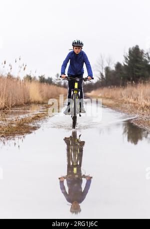 Garçon chevauchant son vélo de montagne à travers une flaque d'eau sur un sentier inondé. Banque D'Images