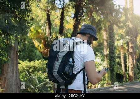 homme touristique barbu millenial portant un chapeau de seau, avec sac à dos et écouteurs à l'aide d'un smartphone pour la navigation Banque D'Images