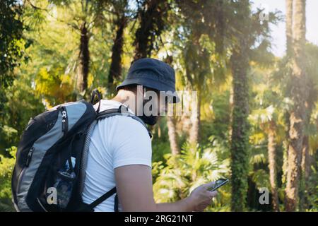 homme touristique barbu millenial portant un chapeau de seau, avec sac à dos et écouteurs à l'aide d'un smartphone pour la navigation Banque D'Images