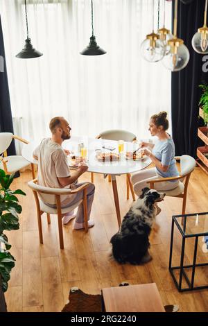 Vue latérale de couple joyeux ayant le petit déjeuner maison près de frontière collie chien à la maison le matin Banque D'Images