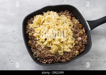 Plat juif traditionnel kasha varnishkes : sarrasin, champignons, oignons frits, pâtes farfalle dans la poêle sur fond gris texturé Banque D'Images