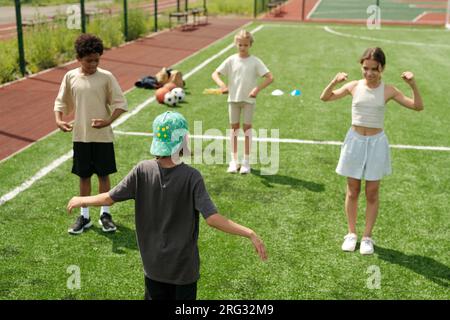 Vue arrière d'un écolier en vêtements actifs debout devant ses camarades de classe et leur montrant de nouveaux exercices physiques pendant qu'ils répètent après lui Banque D'Images