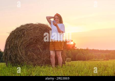 Une femme avec un téléphone portable est debout dans un champ d'herbe tondue avec des piles de foin en arrière-plan contre un coucher de soleil. Banque D'Images