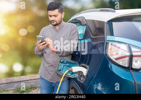 Homme indien utilisant smartphone tandis que sa voiture électrique moderne se charge à la station de charge. Port de recharge de batterie de véhicule électrique. Renouveler Banque D'Images