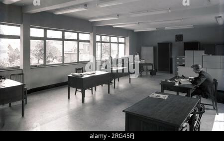 Années 1950, historique, vue d'un bureau ouvert lumineux de l'époque, avec des employés travaillant sur des bureaux individuels en bois, Witney, Oxford, Angleterre, Royaume-Uni. Téléphones Bakerlite, stylo et papier et classeurs. Une dame assise à un bureau utilisant une machine à écrire. Banque D'Images