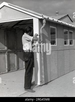 Années 1950, historique, un homme fixant un support sur la gouttière d'un garage préfabriqué, Witney, Oxford, Angleterre, Royaume-Uni. Les garages préfabriqués à auto-assemblage avec des murs en panneaux d'acier/béton sont devenus populaires à cette époque avec l'augmentation de la propriété de voitures à moteur. Banque D'Images