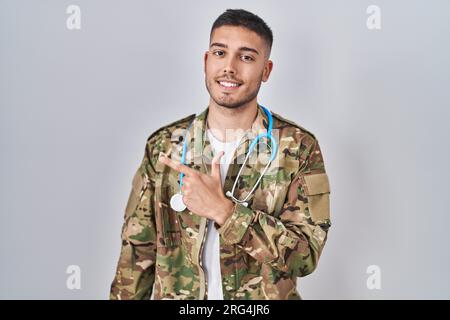 Jeune médecin hispanique portant l'uniforme de l'armée de camouflage souriant joyeux pointant avec la main et le doigt sur le côté Banque D'Images