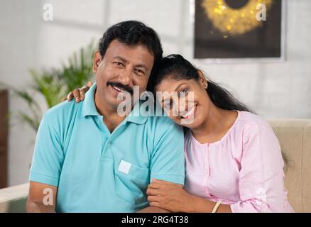 Heureux couple indien d'âge moyen regardant la caméra tout en étant assis sur un canapé à la maison - concept de famille aimante, connexion joyeuse et liaison détendue Banque D'Images