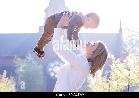 Une scène réconfortante se déroule dans le parc alors qu'une jeune femme blonde millénaire partage des moments de pure joie avec son fils d'un an. Avec un esprit ludique, elle jette avec amour son petit garçon ravi dans les airs, créant un spectacle enchanteur de rire et de bonheur. Le parc devient une toile de bonheur partagé, capturant l’essence d’un lien fort et affectueux entre mère et fils. Joyful Bond : Blonde Millennial Mother lançant de manière ludique son fils d'un an dans les airs au parc. Photo de haute qualité Banque D'Images