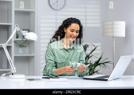 Joyeuse et réussie femme d'affaires au lieu de travail à l'intérieur du bureau à domicile comptant le revenu est arrivé argent dollars américains en espèces. Banque D'Images