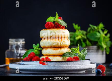 Crêpes épaisses au fromage cottage aux myrtilles, framboises et groseilles rouges, décorées de menthe fraîche sur une assiette grise, délicieux petit déjeuner. Tabl. Noir Banque D'Images