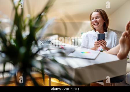 jeune étudiante universitaire femme étudiant utilisant son téléphone portable et riant tout en étant assise au bureau à la maison et en faisant ses devoirs. Banque D'Images