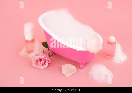 Petite baignoire avec mousse, bouteilles de produits cosmétiques et fleur rose sur fond rose Banque D'Images