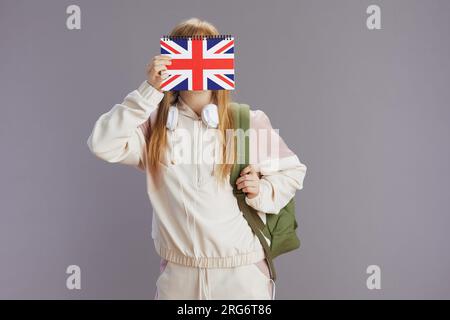 jeune fille moderne en survêtement beige avec sac à dos, cahier de drapeau anglais et écouteurs isolés sur fond gris. Banque D'Images