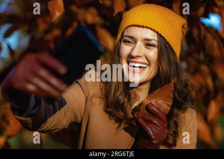 Bonjour novembre. heureuse femme d'âge moyen en manteau beige et chapeau orange ayant la réunion en ligne parmi le feuillage automnal à l'extérieur sur le parc de la ville en automne. Banque D'Images