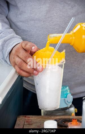 Plan rapproché d'un homme latin versant de la vanille ou du sirop de mangue dans un verre avec de la glace pilée. Snacks que l'on peut trouver dans les rues du Mexique. Banque D'Images