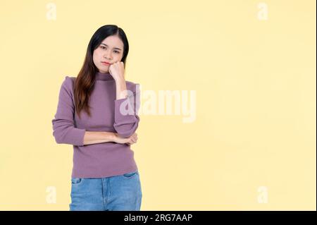 Une femme asiatique ennuyée et bouleversée se tient sur un fond jaune isolé avec sa main sur son menton et regarde la caméra avec un visage malheureux. triste Banque D'Images