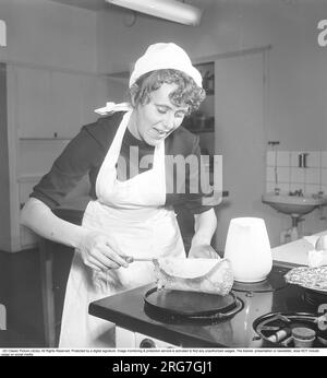 Inger Larsson est debout près d'une cuisinière à gaz et frire des crêpes. Elle retourne une crêpe dans le moule à crêpes en fonte. Sur le côté, elle a une cruche de pâte à crêpes et quelques crêpes qu'elle a déjà faites. Suède 1955. Kristoffersson réf BU76-5 Banque D'Images