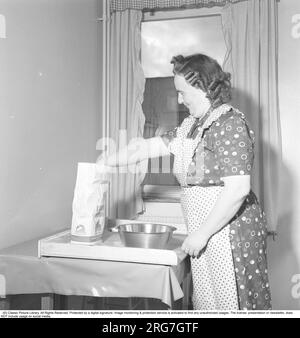 Dans la cuisine dans les années 1950 Une dame âgée regardant très sincèrement à l'aise lorsqu'on la voit cuire sur le plateau en bois spécial qui était une caractéristique commune dans les intérieurs de cuisine des années 1950 et 1960. Elle porte un tablier avec un motif en pointillés. Suède septembre 1952. Banque D'Images