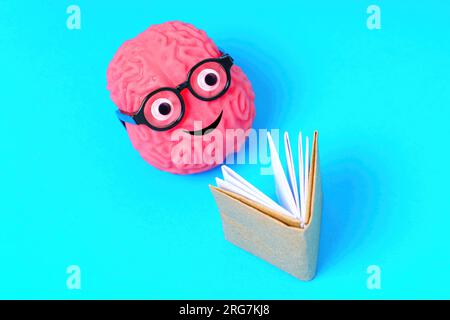 Personnage de cerveau souriant enfilant des lunettes Nerdy, joyeusement absorbé dans la lecture d'un livre, isolé sur fond bleu. Banque D'Images