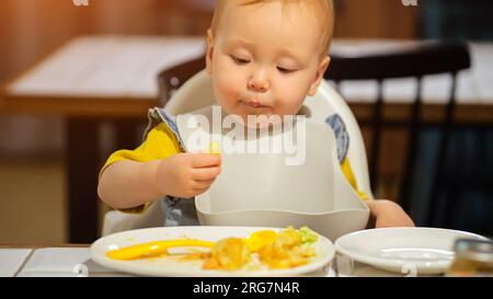 Mignon enfant en bavoir mange un plat avec les mains assises à table Banque D'Images