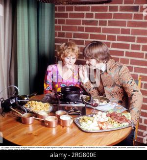 Fondue. Un plat qui a connu sa percée dans les années 60 et 70 Un pot à fondue avec de l'huile chaude se tient au milieu de la table et vous y trempez de la viande pour cuisiner et manger vous-même. Elle porte un chemisier à manches longues très coloré et à motifs qui était typique de la fin des années 60 et 70 Lui dans une veste d'inspiration pop pas sans rappeler les vêtements que les chanteurs portaient à l'époque. Suède 1970. Réf. Conard CV49-5 Banque D'Images