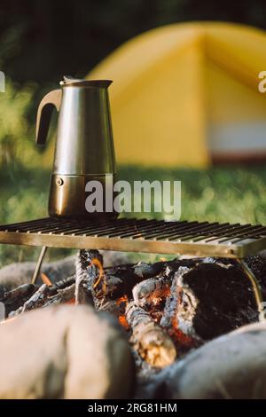 Gros plan de la cafetière geyser sur le feu de joie près de tente dans le parc d'été vert au lever ou au coucher du soleil. Ambiance de camping, nature et style de vie en plein air Banque D'Images