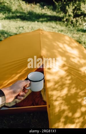 Gros plan d'un jeune homme caucasien tenant une tasse blanche avec du café chaud le matin, profitant de la randonnée. Belles ombres de feuillage d'arbre feuillu reflétées sur le hurlement Banque D'Images