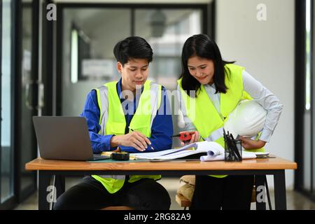 Deux ingénieurs portant des gilets jaunes discutant du nouveau projet en regardant le plan au bureau Banque D'Images