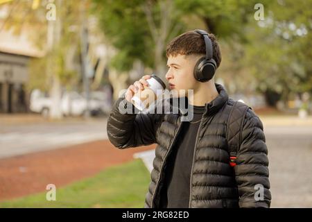 Guy avec des écouteurs boit du café tout en marchant dans un parc public avec espace de copie. Banque D'Images