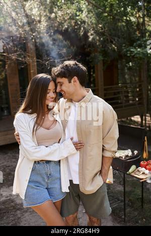 Couple romantique souriant serrant près du barbecue flou et maison de vacances à l'arrière-plan à l'extérieur Banque D'Images