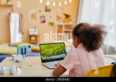 Vue arrière de l'écolière afro-américaine assise par le bureau devant les caméscopes d'ordinateur portable et de smartphone et jouant au jeu vidéo dans l'environnement de la maison Banque D'Images