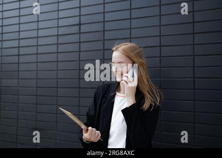 Portrait de femme d'entreprise répond à l'appel téléphonique tout en marchant à l'immeuble de bureaux. Femme d'affaires appelant quelqu'un, tenant des documents. Banque D'Images