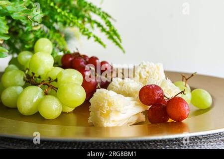Fromage TETE de moine avec raisins verts et rouges sur plaque dorée, décoré de feuilles de fougère, créant un apéritif élégant Banque D'Images