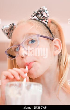 Petite jolie fille de sept ans buvant un milkshake à la fraise à travers une paille dans un café. Portrait de la fille heureuse buvant un cocktail Banque D'Images