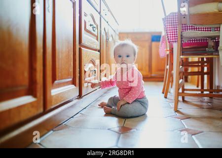 Petite fille assise sur le sol dans la cuisine. Petit enfant rampant à la maison et explorant les choses autour d'elle Banque D'Images