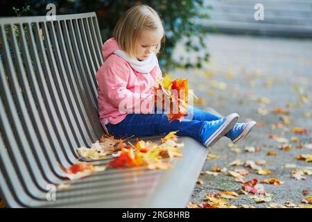 Adorable petite fille assise sur le banc et rassemblant les feuilles tombées dans le parc d'automne. Enfant heureux profitant de la journée d'automne à Paris, France. Activités de plein air Banque D'Images