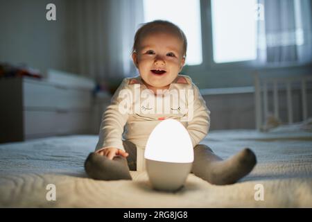 Adorable petite fille jouant avec la lampe de chevet dans la pépinière. Enfant heureux assis sur le lit avec veilleuse. Petit enfant à la maison le soir avant de dormir Banque D'Images