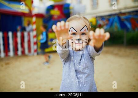 Petite fille d'enfant d'âge préscolaire avec la peinture de visage de chat, faisant grimace drôle à l'extérieur. Peinture de visage d'enfants. Activités créatives pour les enfants Banque D'Images