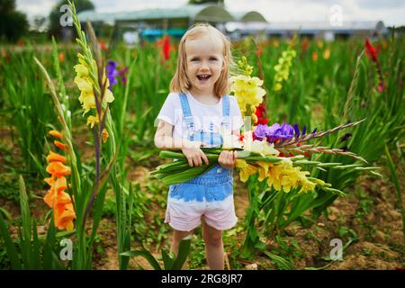 Adorable fille cueillant de belles fleurs de gladiolus à la ferme. Activités estivales en plein air pour les petits enfants. Banque D'Images