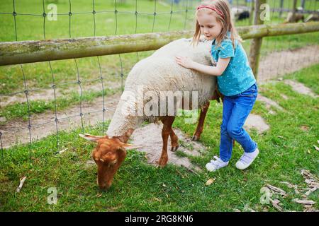 Adorable petite fille jouant avec des chèvres et des moutons à la ferme. Enfant se familiarisant avec les animaux. Agriculture et jardinage pour les petits enfants. Extérieur Banque D'Images