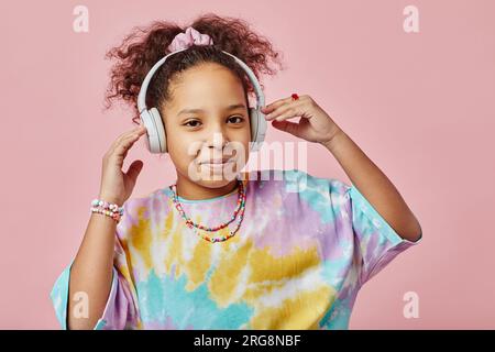 Mignonne écolière en vêtements décontractés touchant des écouteurs et écoutant de la musique tout en se tenant devant la caméra sur fond rose dans le studio Banque D'Images