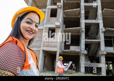 Portrait de souriant jeune femme indienne ingénieur civil portant casque et gilet debout sur le chantier de construction. Immobilier. Regarder la caméra Banque D'Images