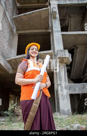 Portrait de jeune confiant belle indienne ingénieur civil ou architecte portant casque et gilet tenant le plan de paperasse à la construction si Banque D'Images