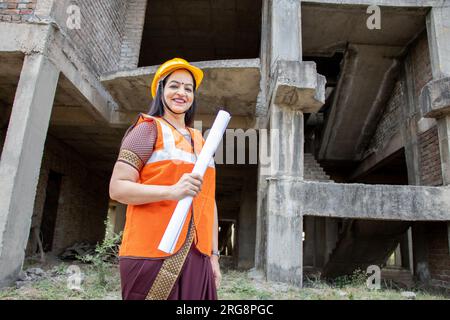 Portrait de jeune confiant belle indienne ingénieur civil ou architecte portant casque et gilet tenant le plan de paperasse à la construction si Banque D'Images