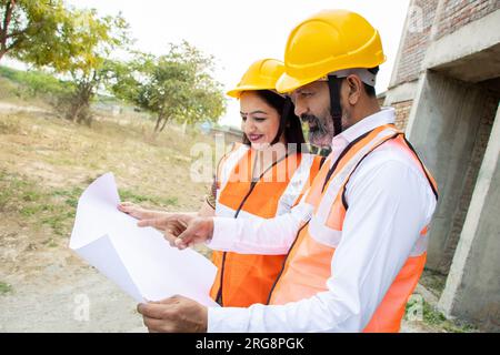 Deux ingénieurs civils indiens hommes et femmes ou un architecte portant casque et gilet tenant le plan de paperasse sur le chantier de construction discutant du vrai estat Banque D'Images